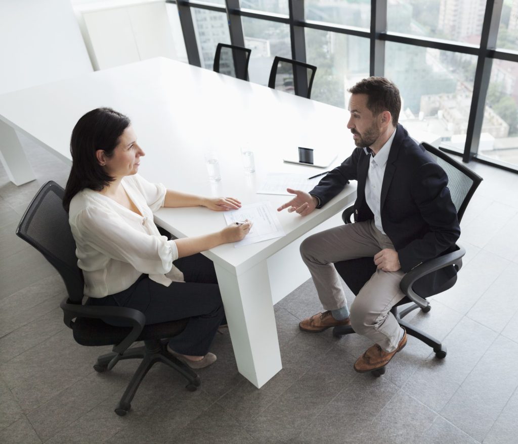 Two people converse over a white table in an open office space